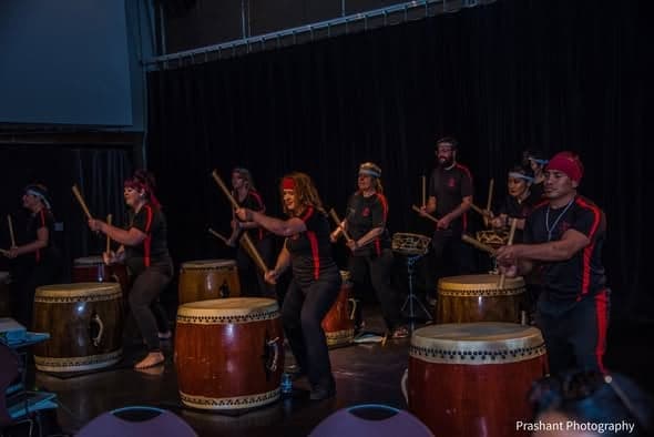 A taiko drumming group dressed in black and red strike large barrel drums on stage.