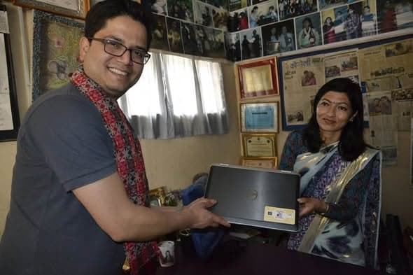 A man in a Nepali scarf hands a laptop across a desk to a woman in a sari, in an office whose walls are covered with photographs, certificates and press cuttings.