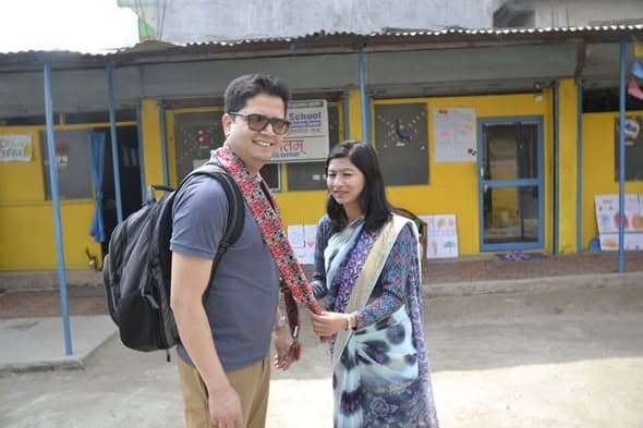 A man with a rucksack and a woman in a sari stand in the yard of a small yellow and blue school building hung with children's drawings.