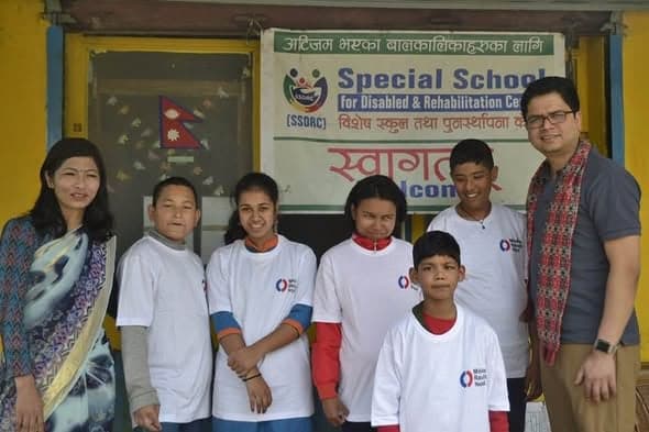 Five students in Mission Rebuild Nepal t-shirts standing with two adults under a painted SSDRC welcome banner at the school entrance.