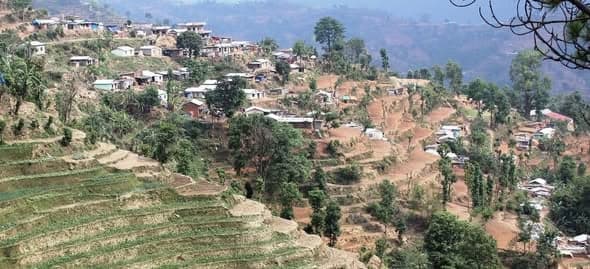A steep hillside scattered with metal-roofed houses among terraced fields and bare red earth.