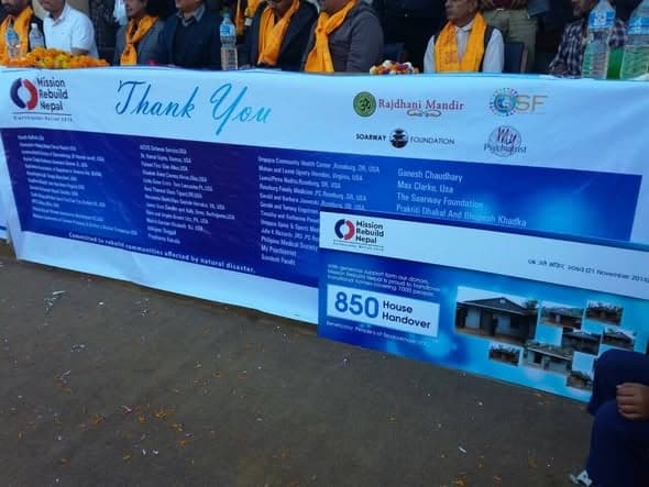 A long printed banner reading Thank You and 850 House Handover, listing donor organisations, hung behind a table of guests at an outdoor ceremony