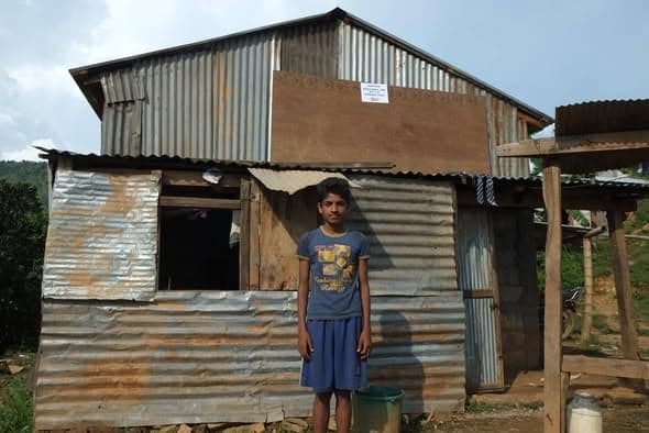 A boy in a blue T-shirt stands in front of a two-level corrugated-iron house built on a stone base.