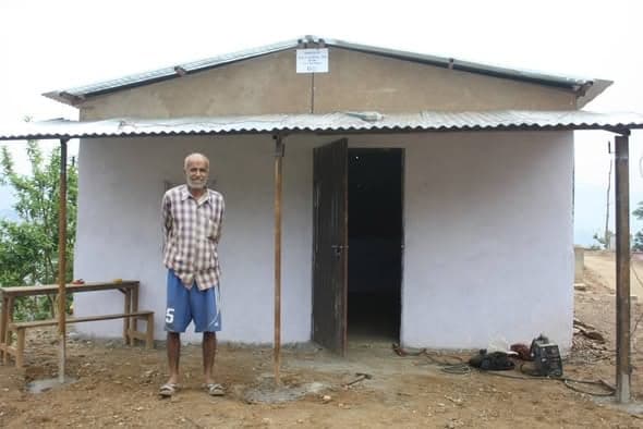 An older man stands under the eaves of a newly whitewashed house with an open doorway and tools left on the ground.