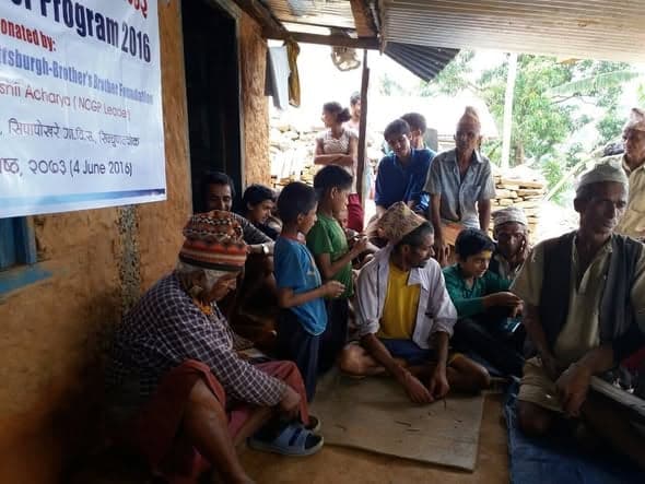 Villagers of all ages sit and stand under the eaves of a house at a handover gathering, beside a printed programme banner.