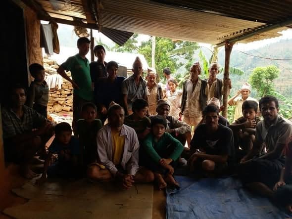 A group of men, women and children sit together on mats and tarpaulins under a corrugated roof, looking towards the camera.