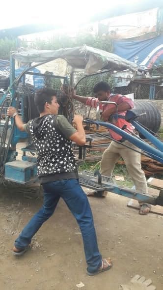 Two young men load steel reinforcing bar onto a small blue tractor trailer in a roadside yard.