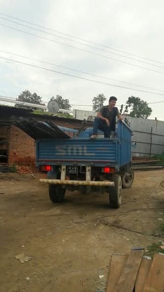 A man sits on the rail of a loaded blue tipper truck stacked with corrugated roofing sheets.