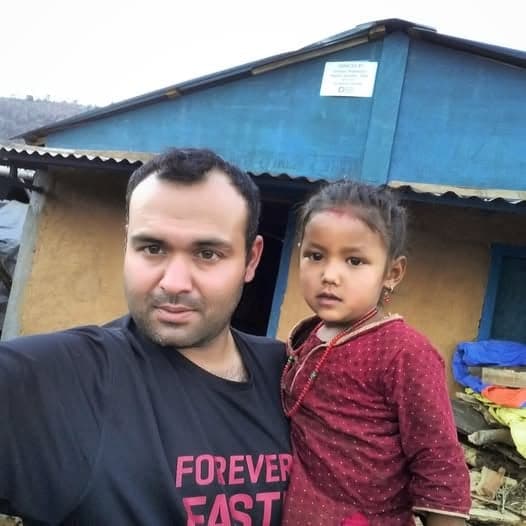 A man holds a young girl on his hip in front of a small blue-painted house on a hillside.