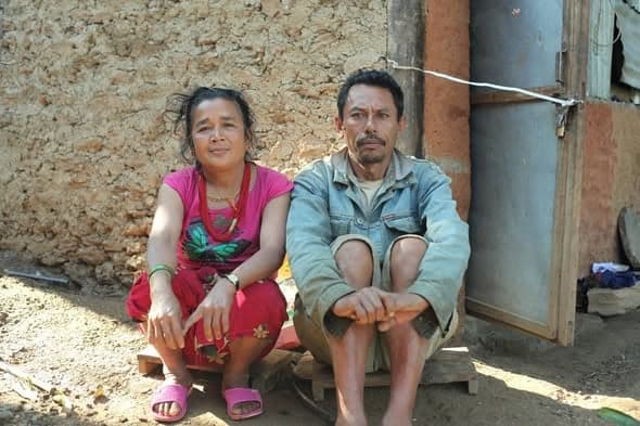 A woman and a man sit on low wooden boards against a mud wall, resting between tasks.