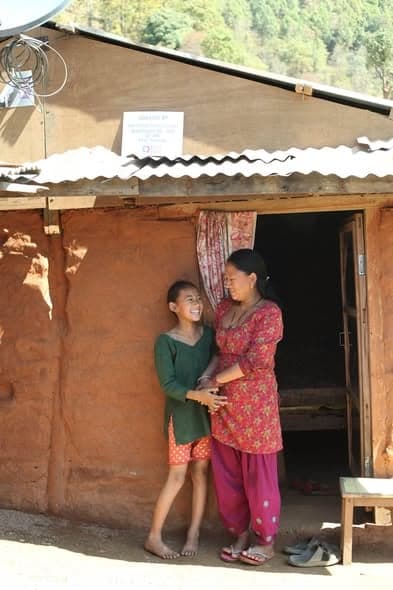 A girl leans against a woman in the doorway of a mud house, both smiling, a sleeping platform visible inside.