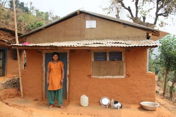 A woman in an orange kurta stands in the doorway of a mud house with cooking vessels set out on the ground outside.