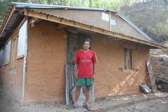 A man in a red football shirt and topi stands against the wall of a newly plastered mud house on a slope.
