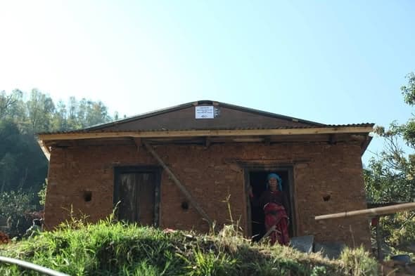 A woman in red stands in the doorway of a newly built mud-brick house against a bright sky.