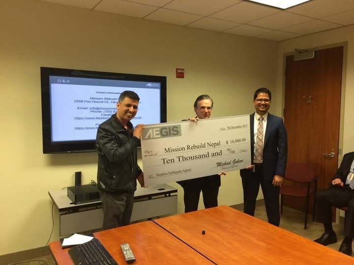 Three men hold an oversized cheque for ten thousand dollars made out to Mission Rebuild Nepal in a meeting room, a presentation on the screen behind them.