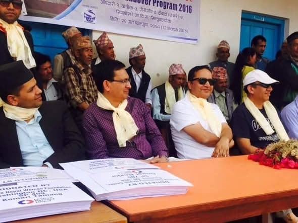 Guests in white scarves sit at a table stacked with printed documents beneath a House Handover Program 2016 banner.