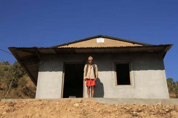 An elderly man stands in the doorway of a newly rendered house with a corrugated roof against a deep blue sky.