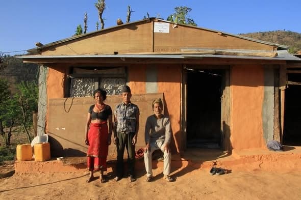 Three people stand and sit in front of a mud house lit by low evening sun, sandals left at the step.