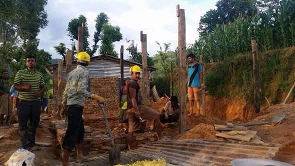 A construction crew in yellow helmets works among timber posts and cut stone on a hillside site.