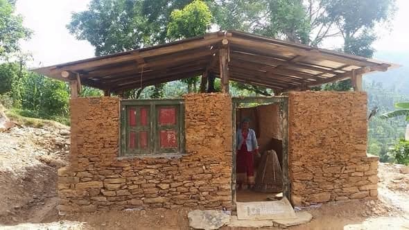 A woman stands in the doorway of a finished stone and mud house with red-painted shutters and a corrugated roof.