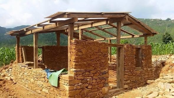 A stone house shell with a completed timber roof frame and corrugated sheets, standing on a terrace above maize fields.