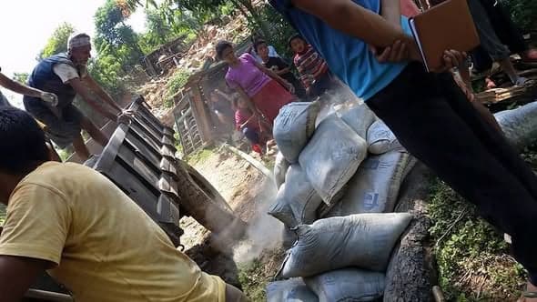 People unload sacks of cement from a truck bed beside a village path while children watch.