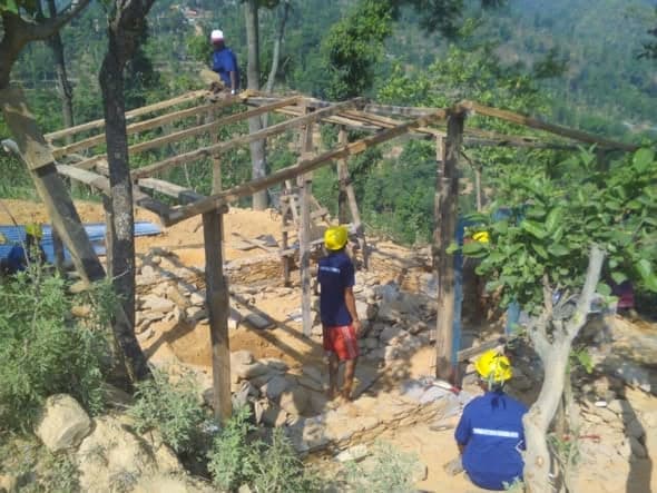 Workers in helmets raise the timber roof frame of a house over newly built stone walls.