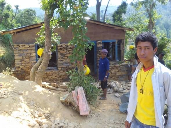 A young man stands in the foreground while workers finish a stone house with a corrugated roof behind him.