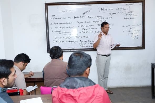 A man in a striped shirt stands at a whiteboard headed Mission Critical, working through a list of tasks with a seated group.