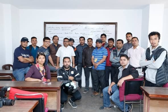 About eighteen volunteers pose for a group photograph in front of the planning whiteboard, camera equipment on the desks.
