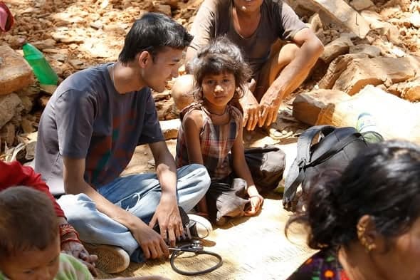 A young man crouches with a blood pressure cuff beside a small girl and her family on a mat spread over rubble.