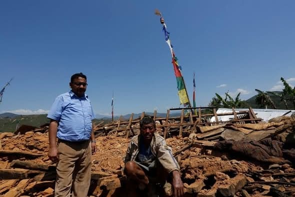 A man in a blue shirt stands with a villager crouching on the rubble of his house, prayer flags flying above.