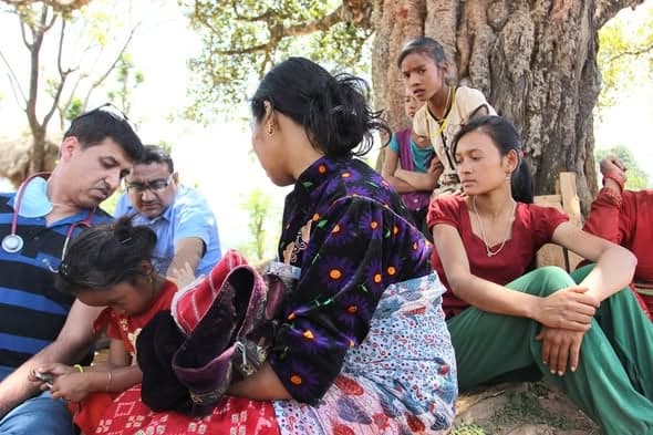 Two doctors, one with a stethoscope, examine a girl lying on a blanket under a tree while her family sits beside her.