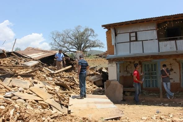 People stand among rubble beside a badly damaged two-storey house, its ground floor wall split open.