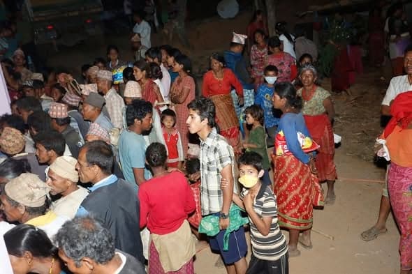 A large crowd of villagers, many with children, wait in line at dusk behind a rope barrier.