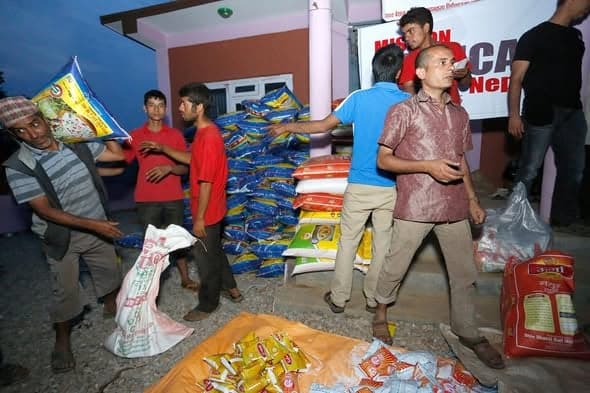 Volunteers pass sacks of rice and packets of food from a large stack inside a building, a Mission Critical Nepal banner on the wall.