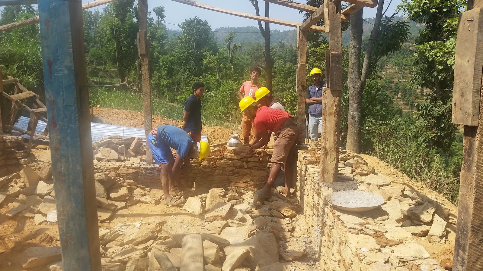 Masons in yellow helmets lay flat stone into a wall while others watch from the edge of the site.