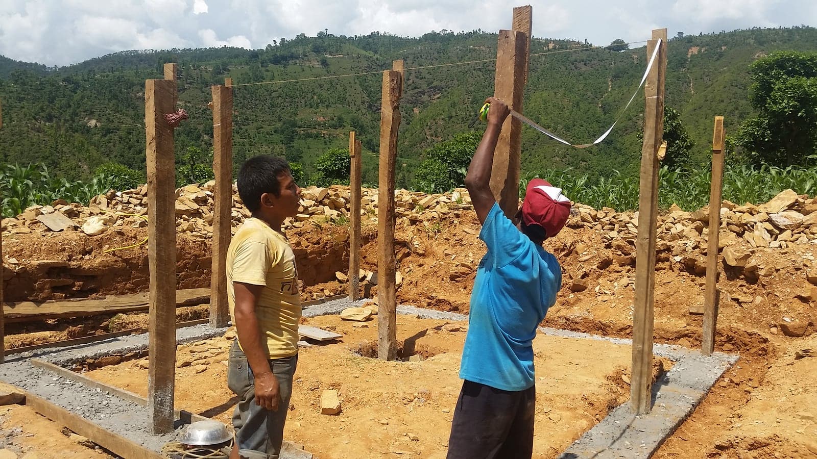 Two men set out timber posts on a new concrete footing, one holding a tape measure against an upright while the other watches.