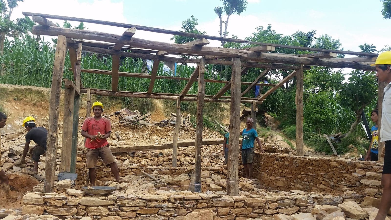 A crew in hard hats builds dry-stone walls beneath a completed timber roof frame, rubble from the collapsed house heaped around them.