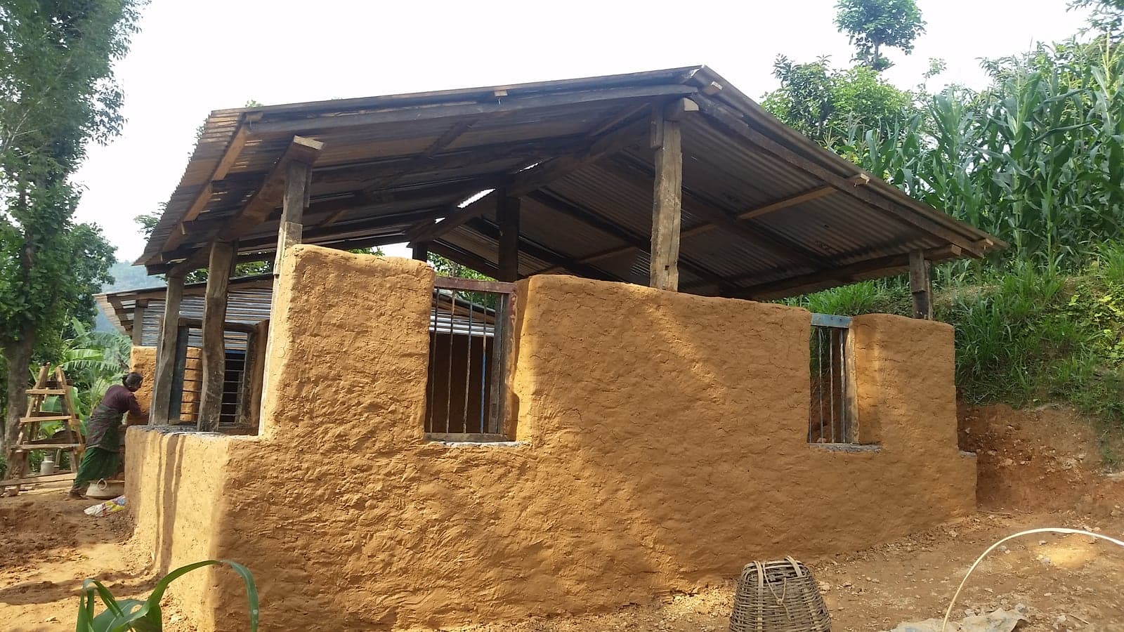 A nearly finished house with smooth mud-rendered walls and barred windows under a deep timber roof; a woman works at the far wall.