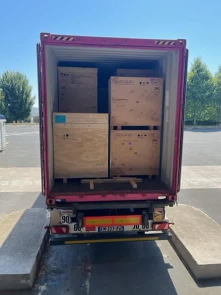 A shipping container loaded with large crated equipment on wooden pallets, photographed from behind an open truck
