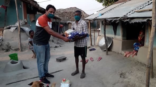 Two masked men handing over a bundle of food supplies in the courtyard of a mud-walled house
