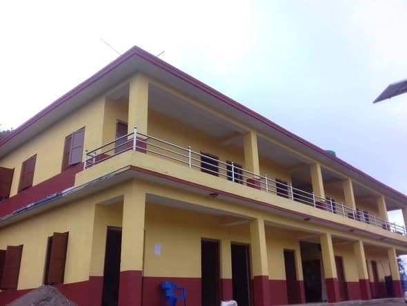A finished two-storey school building painted yellow with a maroon plinth and roofline, with open verandas and steel balustrades on both floors.