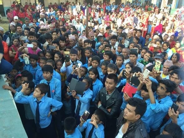 A dense crowd of schoolchildren in blue uniforms and villagers holding up phones to photograph something off-camera