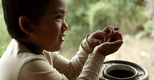 A boy in a pale shirt cupping his hands, filmed in soft daylight beside a metal water vessel