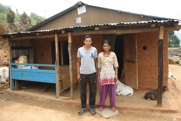 A man and a woman standing in front of a completed transition home with a corrugated roof, a mud-plastered front wall and a blue-painted verandah bench