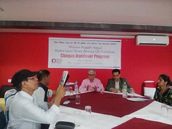 People seated around a conference table beneath a printed banner reading Mission Rebuild Nepal and Rudra Gauri Shree Ambika Oli Pratisthan Cheque Handover Program