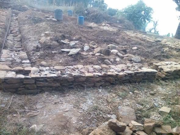 A dry stone plinth built into a cut terrace on a hillside, with blue water drums standing at the top of the slope