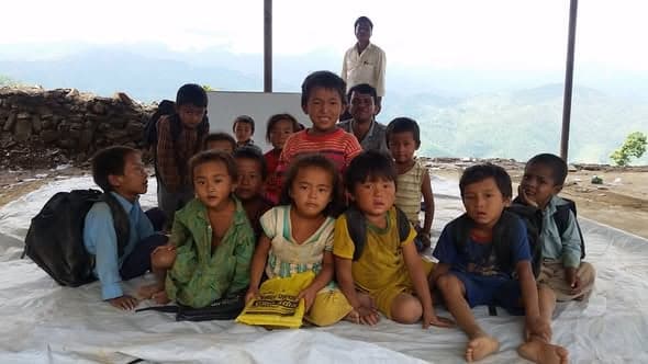 A group of small children sitting on a tarpaulin under an open shelter with a portable blackboard, mountains behind them