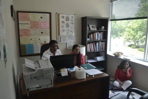 Volunteers working at a desk with a computer and printer in a small clinical office, medical charts on the wall behind them